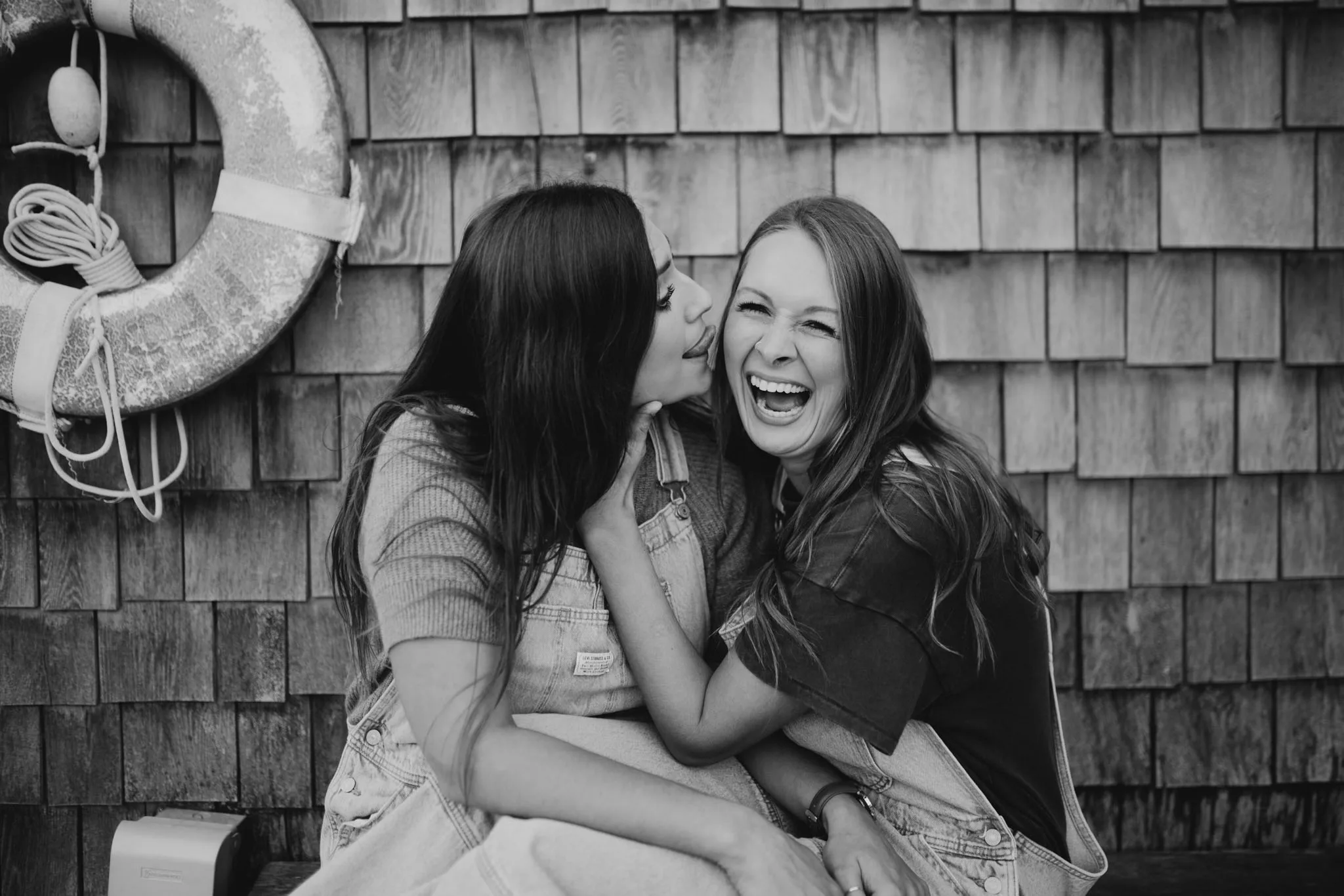 Black and white photo of McKenzie and Kayla laughing together against a cedar shingle wall at Leland Marina