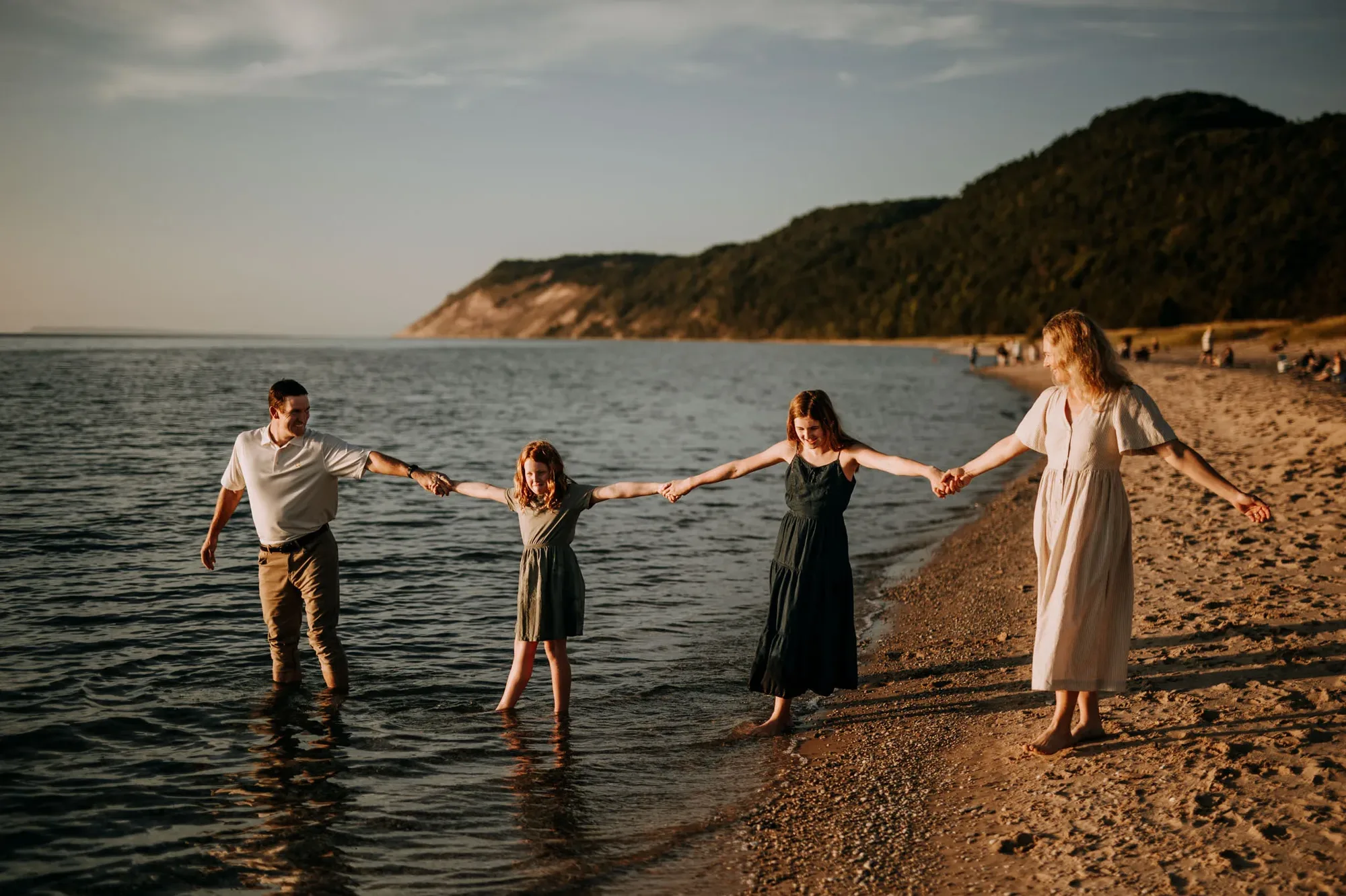 A family of four holding hands in a line, ankle-deep in Lake Michigan at Esch Beach, the bluff behind them