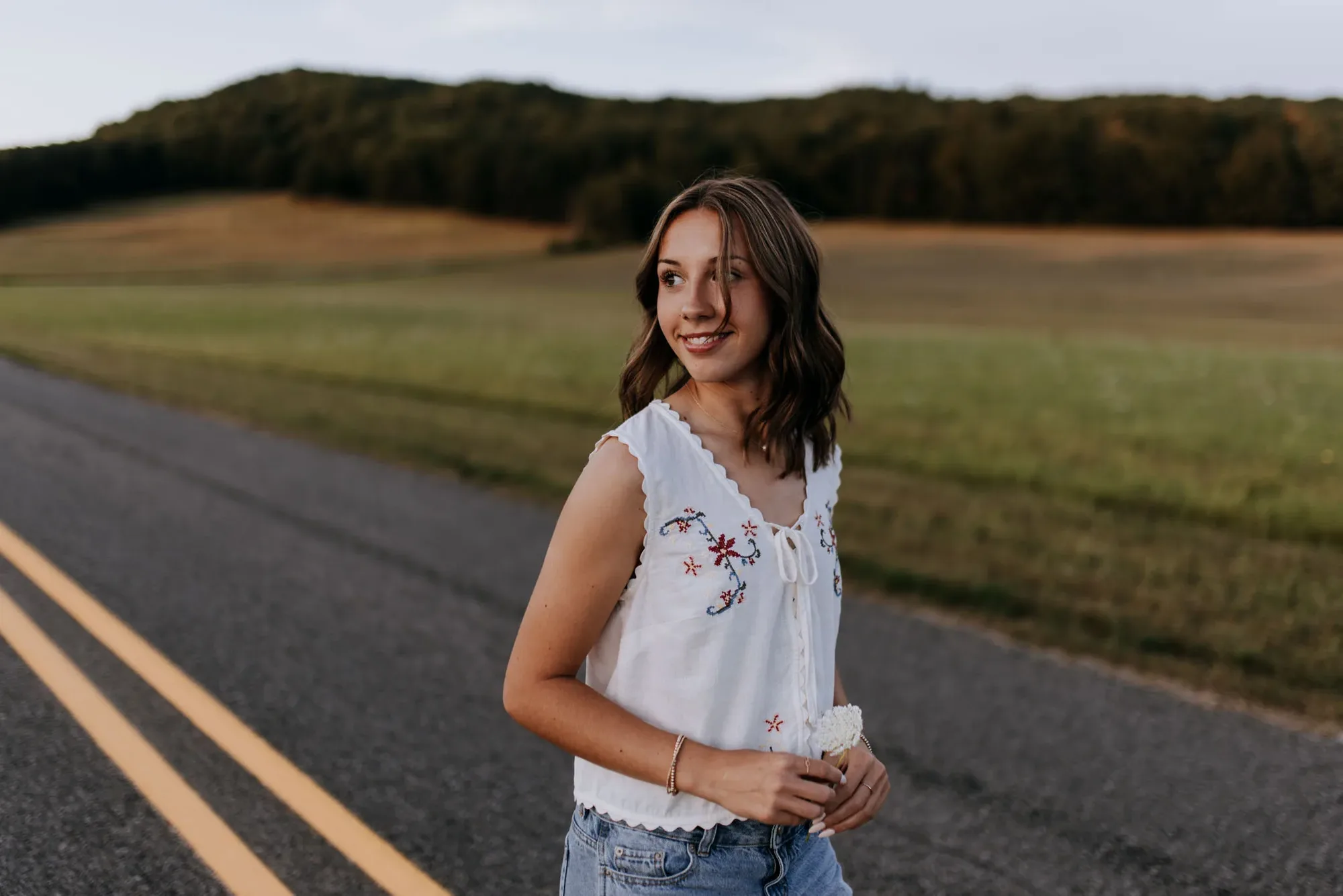 A senior in a white embroidered top standing on a country road at dusk, looking off to the side, a green field and treeline behind her