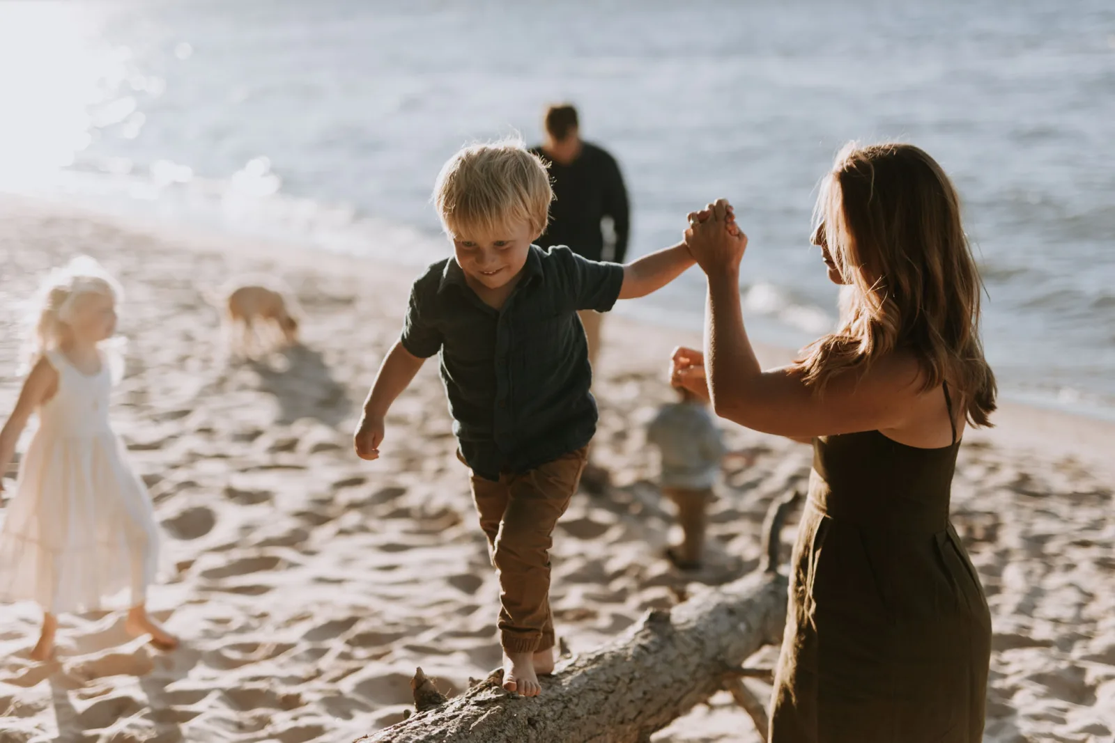 A young boy in a dark shirt walks along a driftwood log while holding hands with a woman on a rocky beach during golden hour.