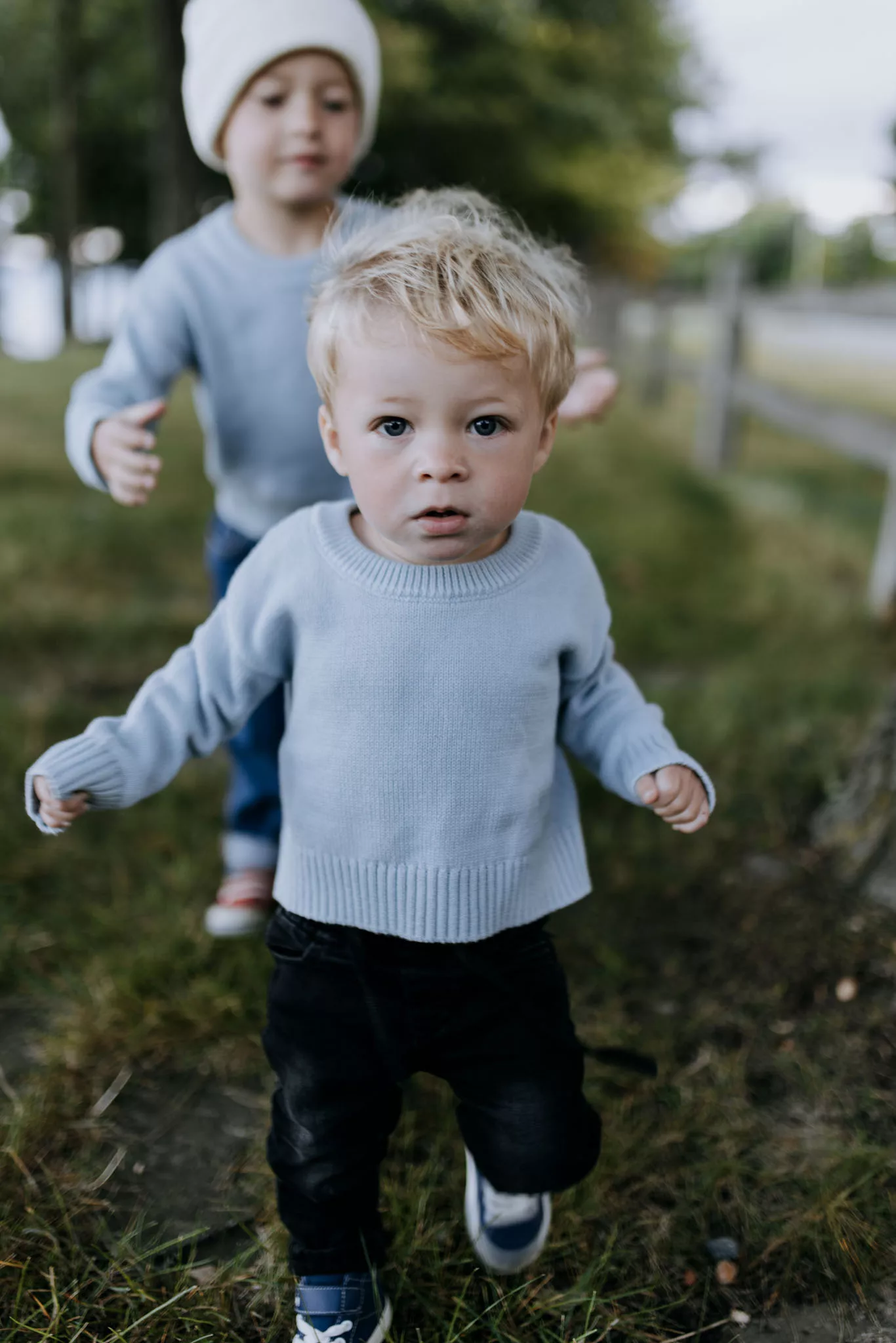 A blonde toddler in a light blue sweater runs through grass with a white-hatted child following behind in an outdoor garden setting.