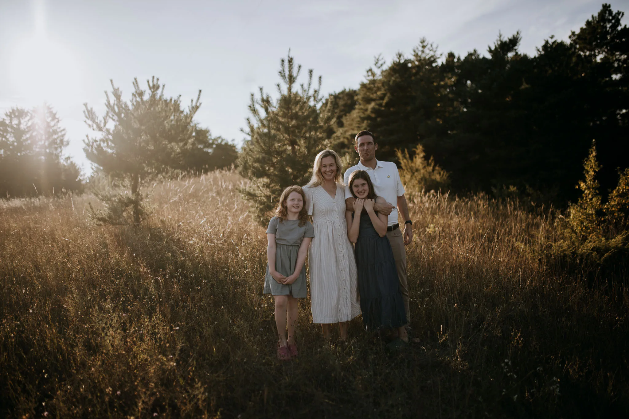 A family of four stands smiling in a field of dried grasses with evergreen trees behind them, bathed in warm golden sunlight.
