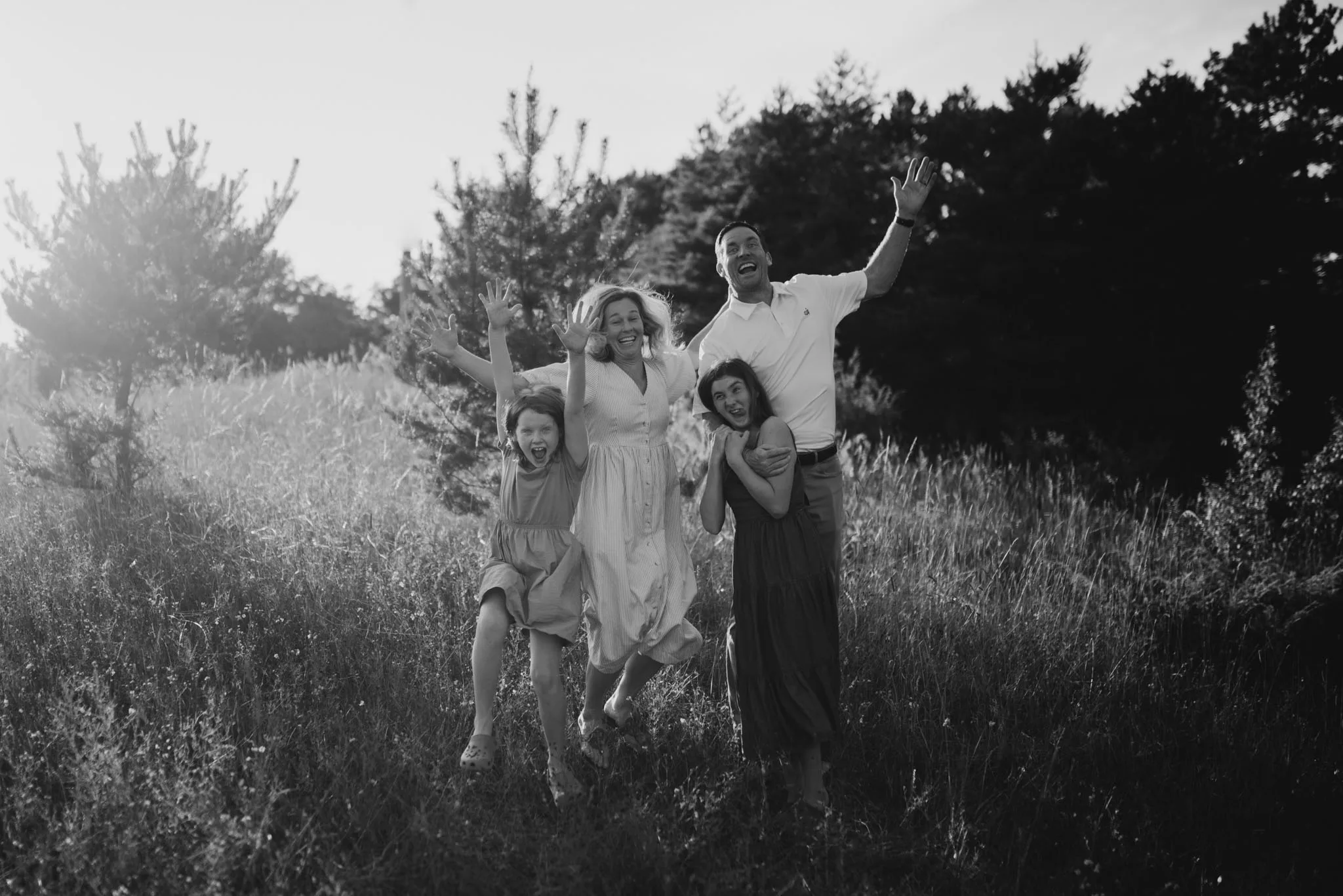 A family of four people jumping with arms raised in celebration in a wildflower field with trees in the background, captured in black and white photography.
