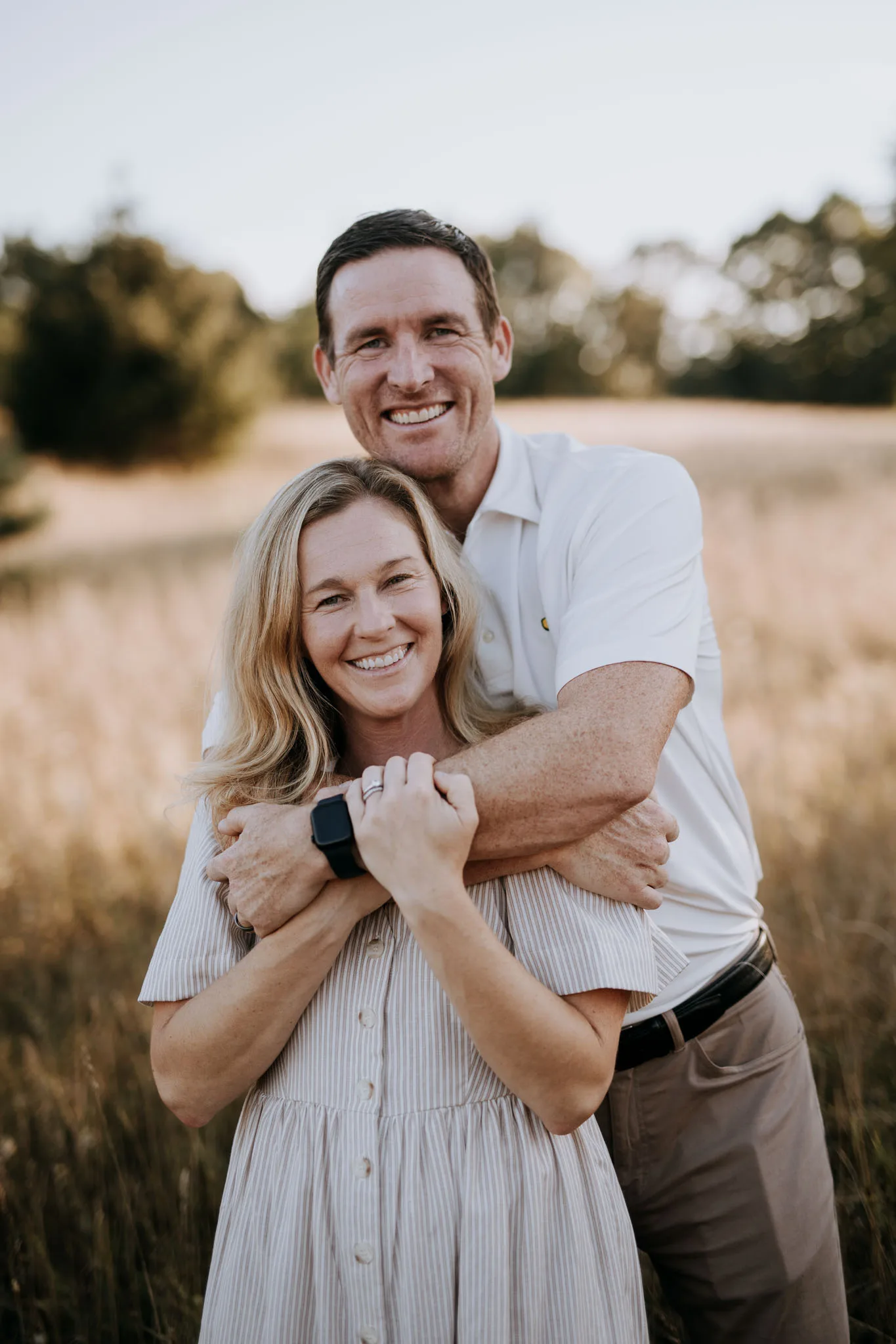 A smiling couple embraces in a golden field, with the man in a white polo shirt and the woman in a light striped dress, surrounded by trees and clear sky.
