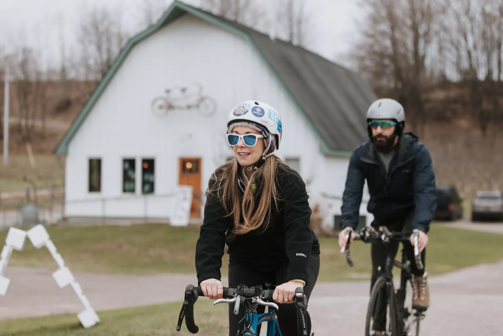 Tiffa and Karl riding bicycles in front of the white barn at Tandem Ciders after the proposal
