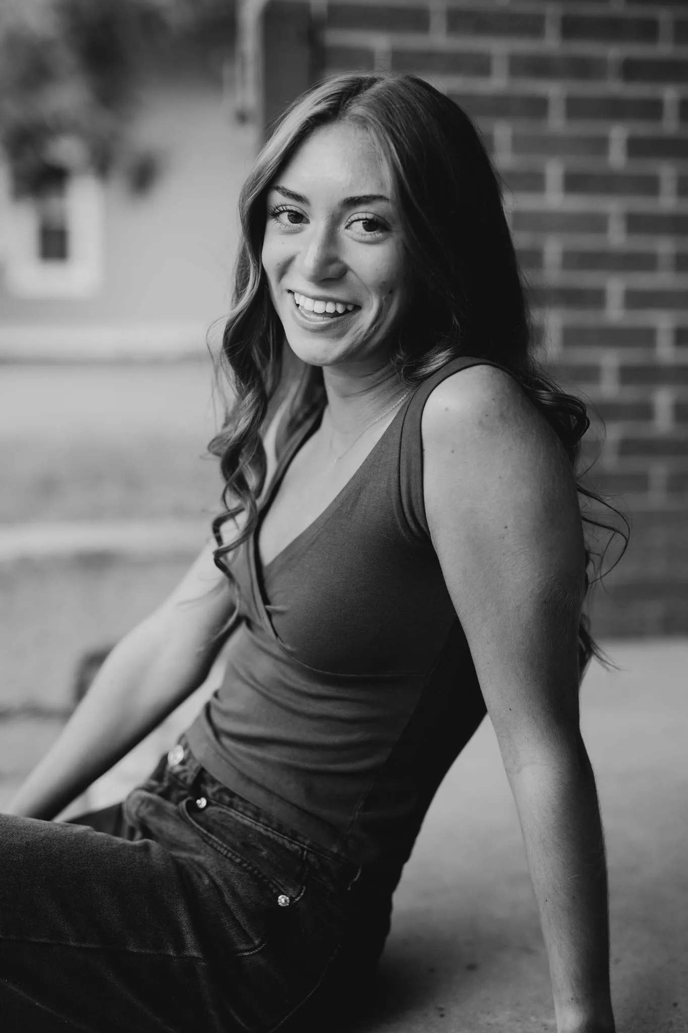 A smiling young woman with long dark hair wearing a sleeveless top sits against a brick wall in this black and white portrait photograph.