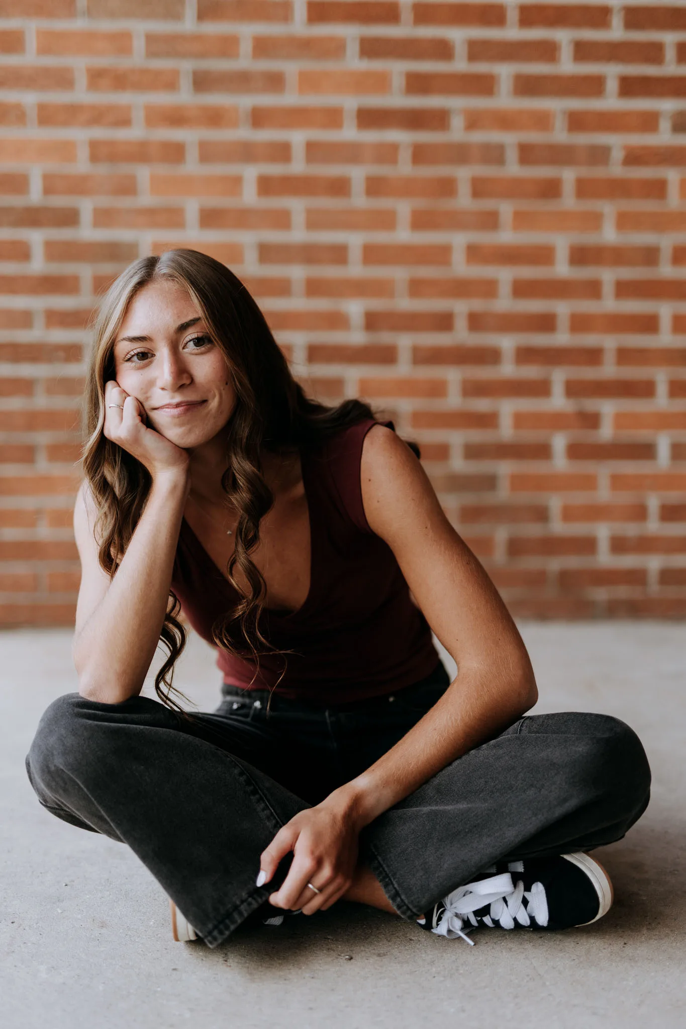 A young woman with long brown hair sits cross-legged against a brick wall wearing a burgundy sleeveless top and dark jeans, resting her chin on her hand with a gentle smile.