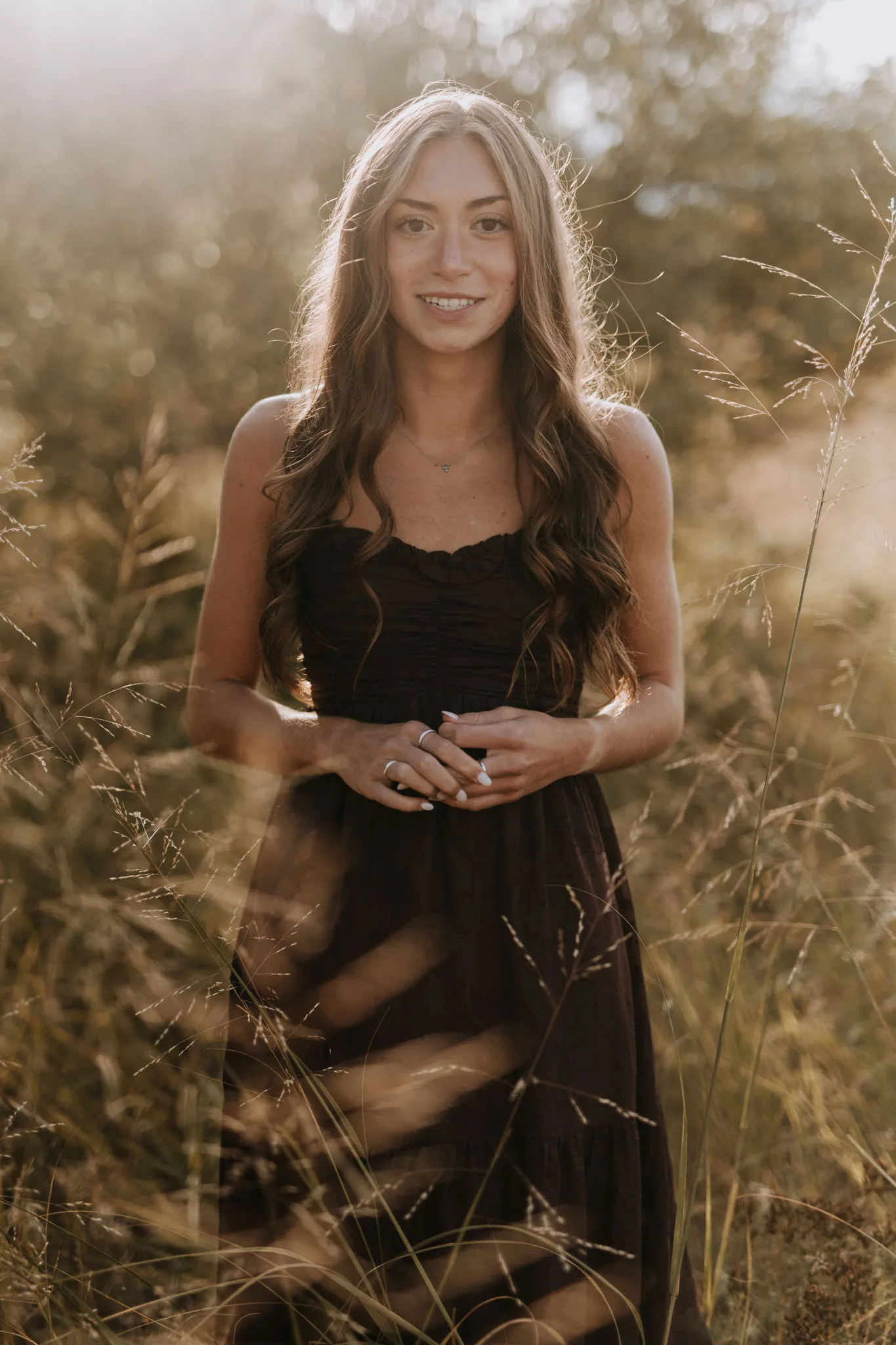 A smiling young woman with long dark hair in a black dress stands among golden dried grasses and wildflowers backlit by warm sunset light.