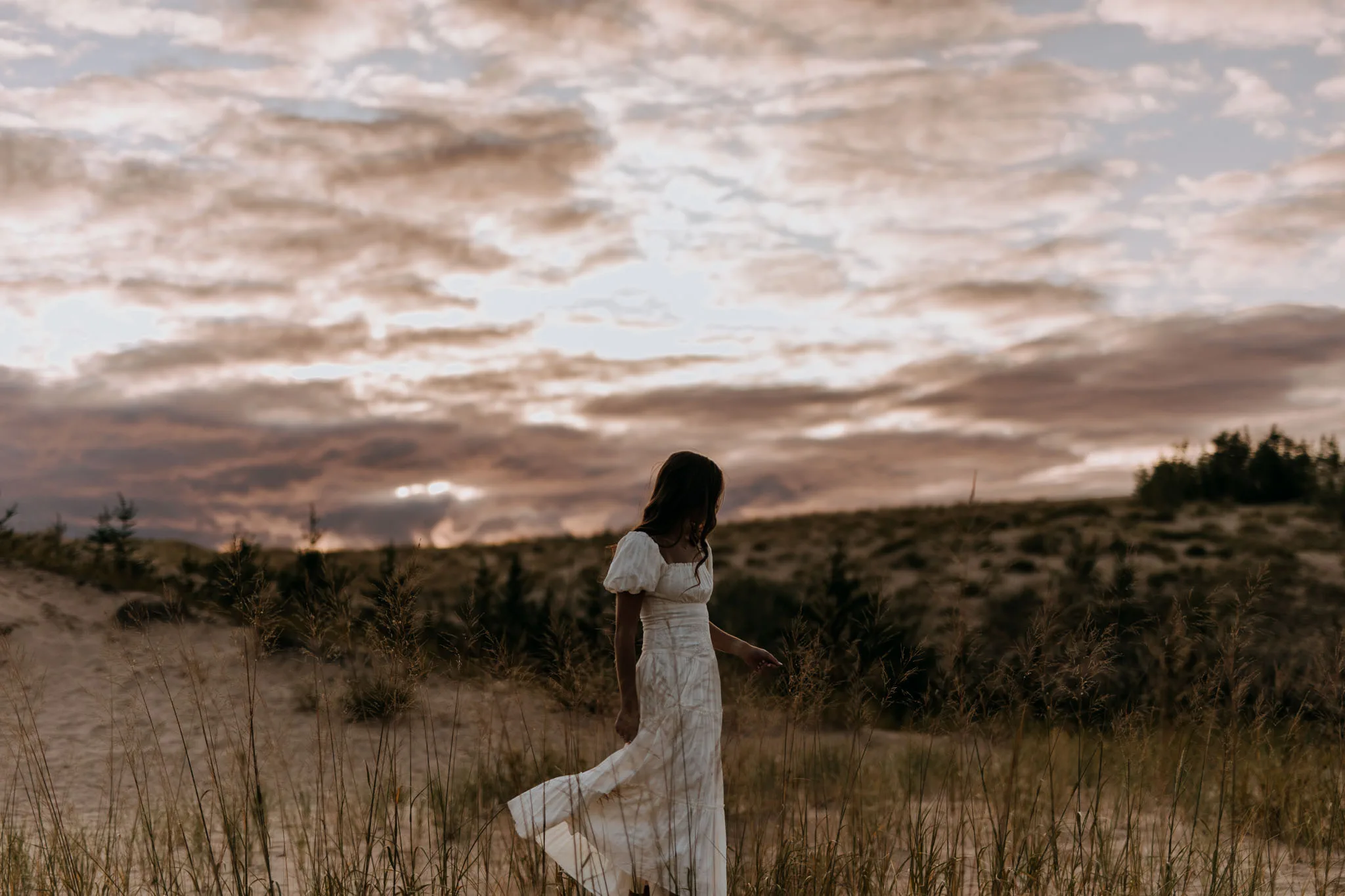 A woman in a white dress stands in a windswept field with evergreens, looking toward distant hills under a dramatic cloudy sky.
