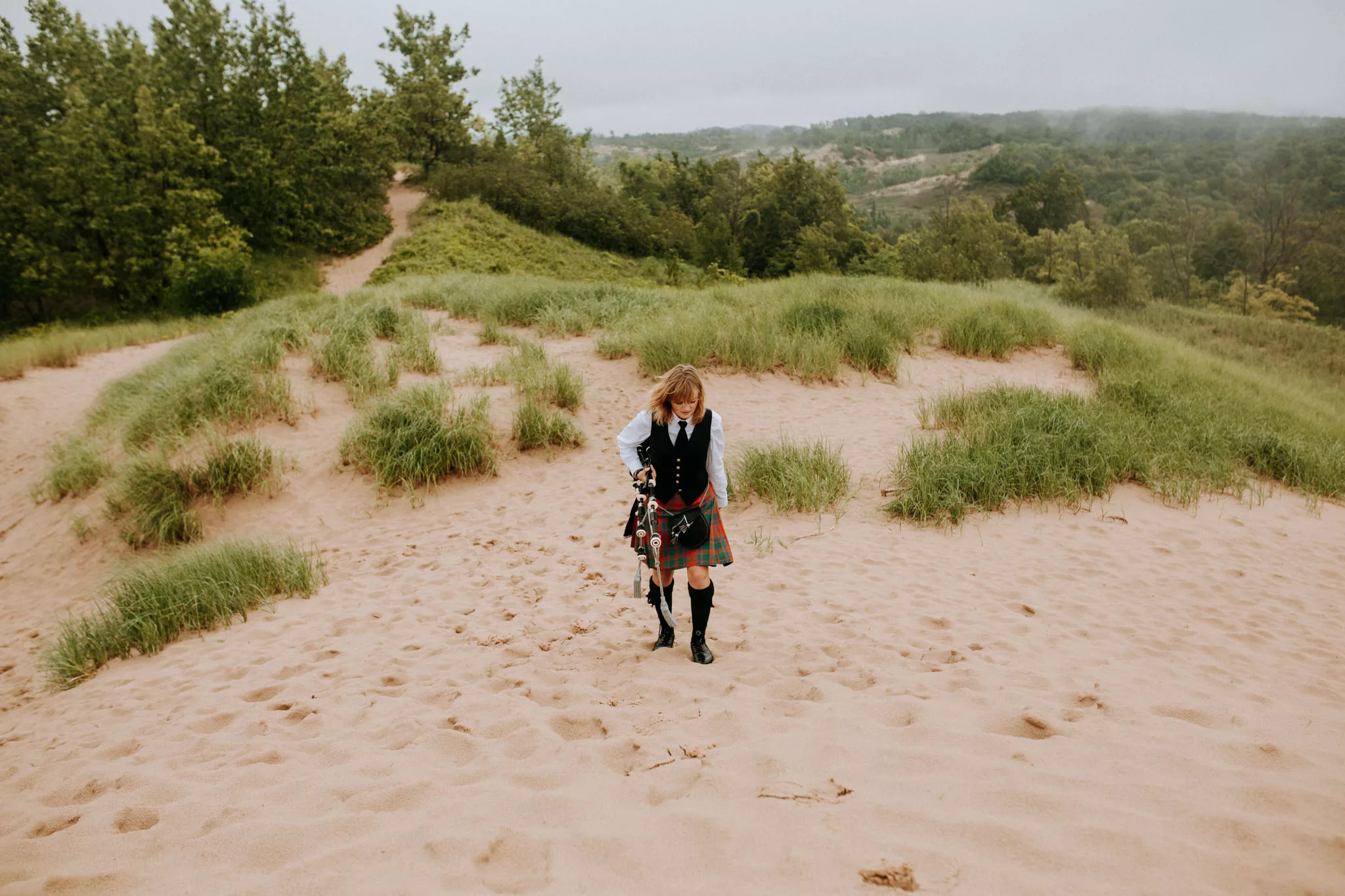 A woman in Scottish traditional dress walks across sandy beach dunes carrying bagpipes with forested hills visible in the misty background.