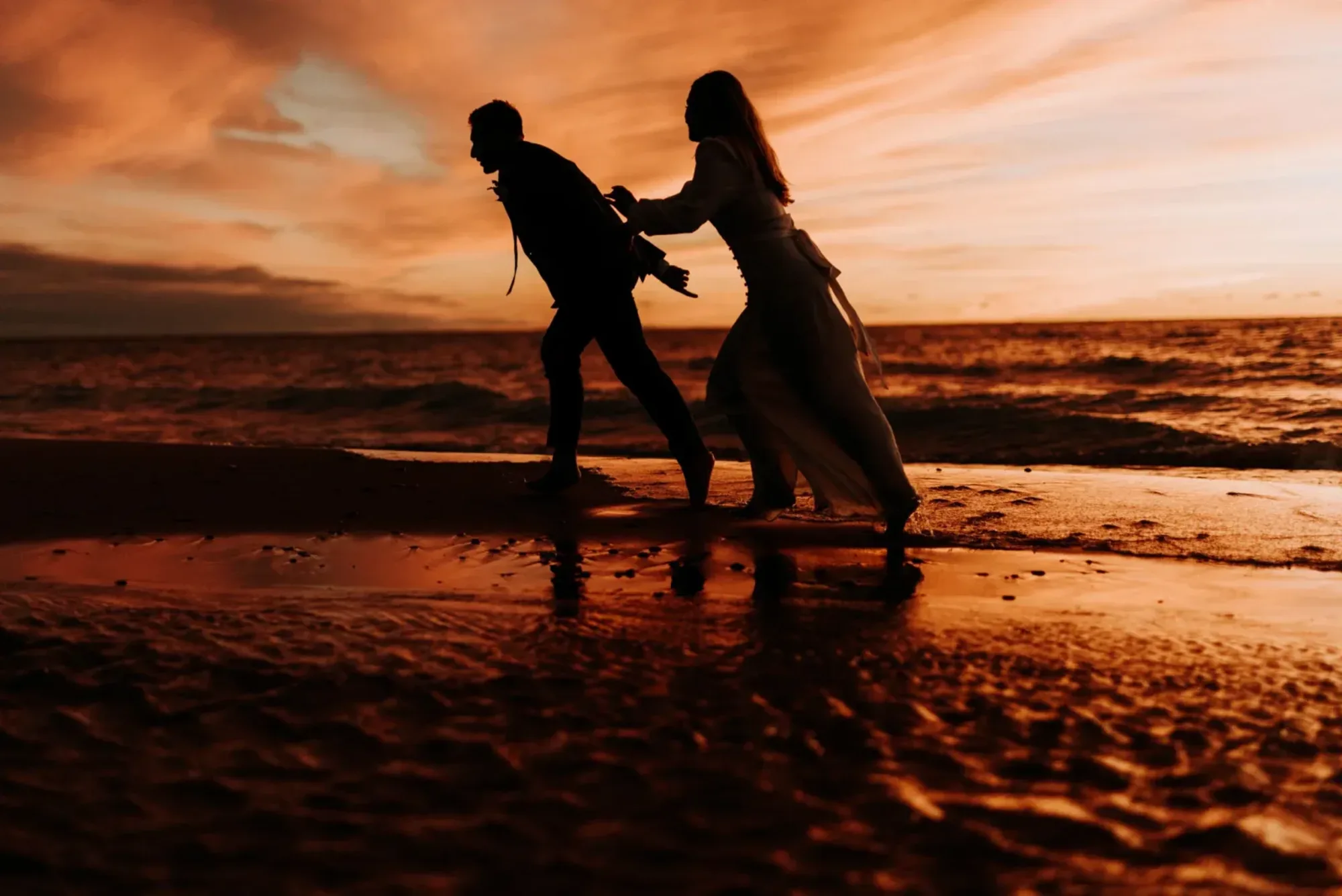 A couple running along the water's edge at sunset, in silhouette against an orange sky, the sand glowing behind them