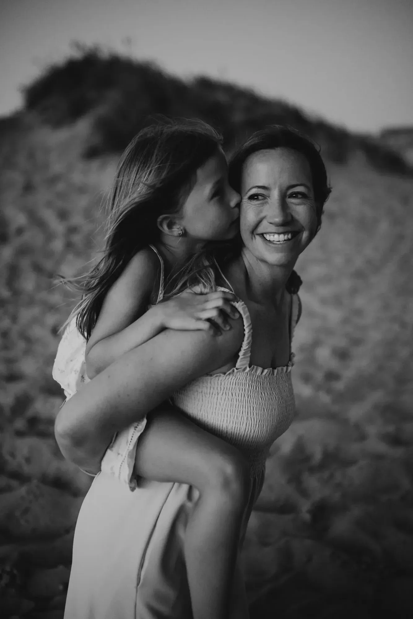 A mom carrying her daughter on her back on the dunes, the girl kissing her cheek, in black and white