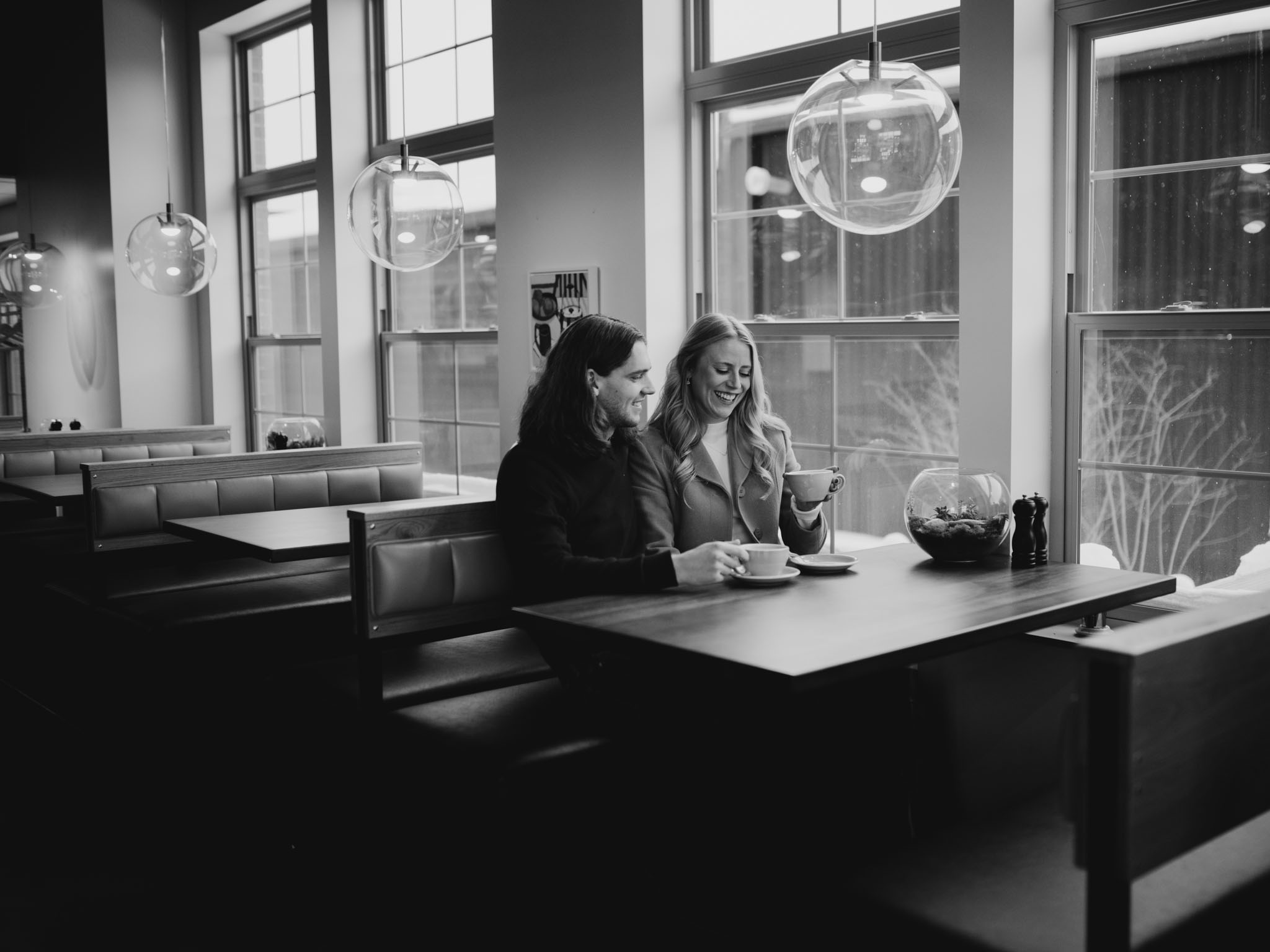 Couple sitting close together at a coffee shop window during winter engagement session warm-up
